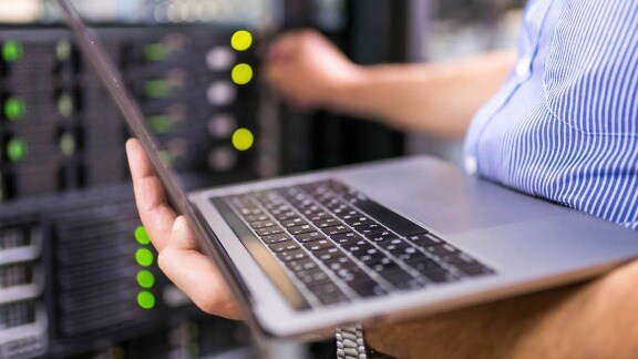 A man stands in front of a server rack, holding a laptop