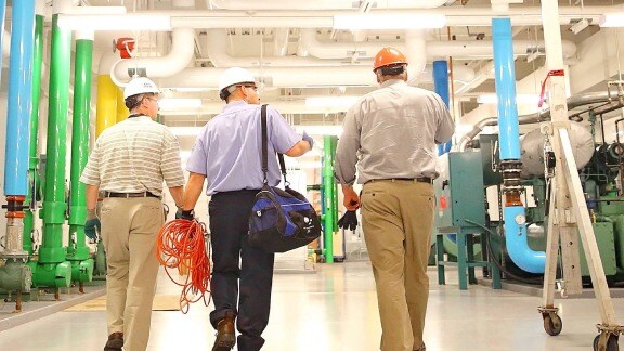 In a factory, few men walk amidst green pipes, illustrating the industrial atmosphere of the space