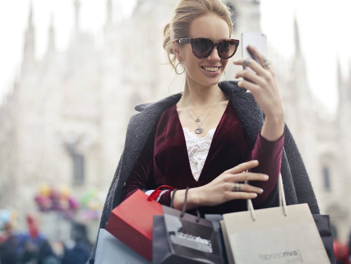 A woman wearing sunglasses captures a selfie, surrounded by shopping bags in a vibrant setting.