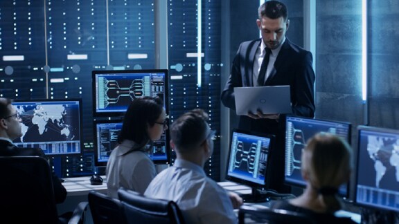 A employee holding the laptop in hand and heading the discussion with the others in the cybersecurity center